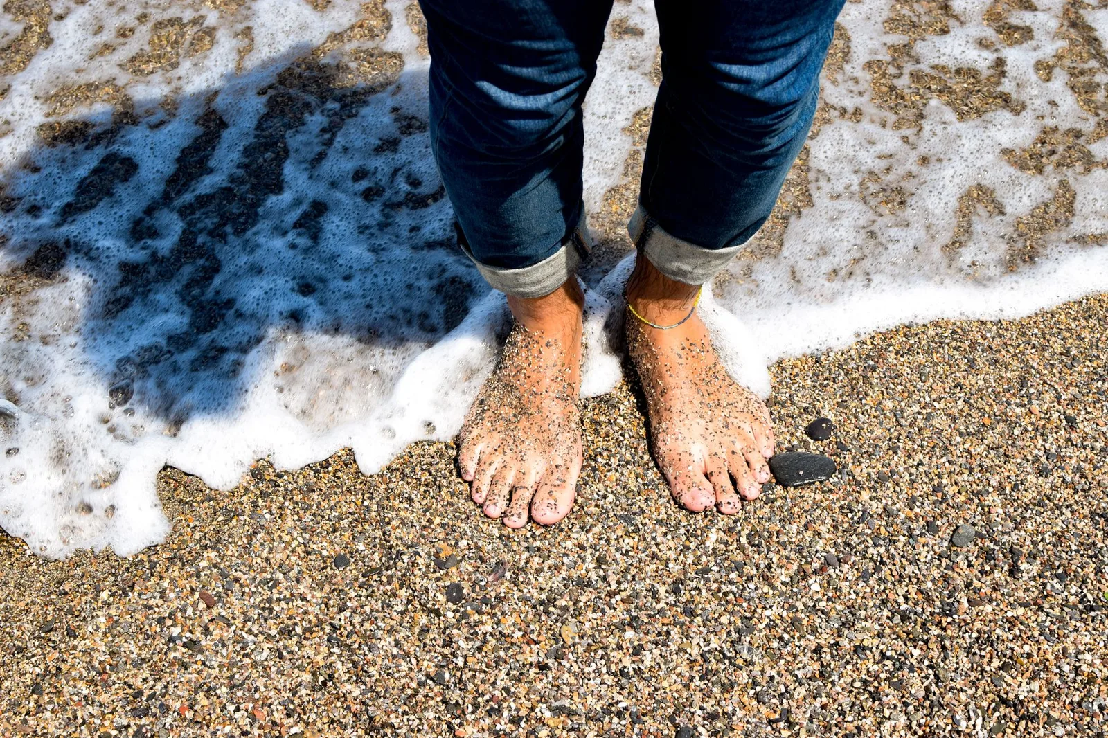 Feet in jeans standing on a sandy beach with waves lapping. Perfect summer day at the seaside.