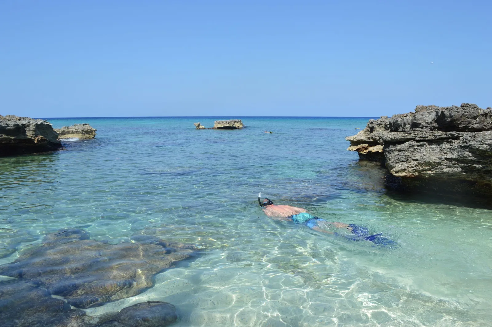 A person snorkeling in crystal clear tropical waters surrounded by rocky formations and a vast serene ocean.