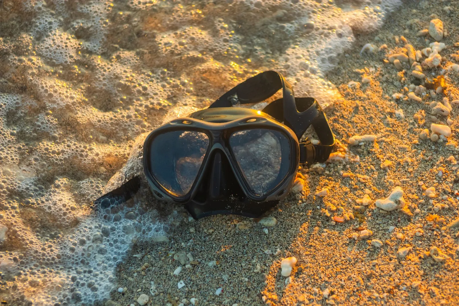 A scuba mask rests on a sandy shoreline as waves gently wash over it during sunset.
